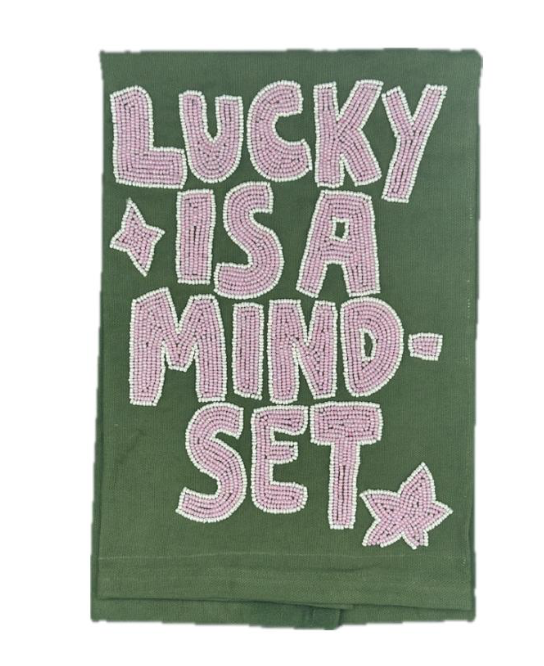 A dark green tea towel featuring the phrase 'LUCKY IS A MIND-SET' in large, pink and white textured letters, accented with small pink stars.