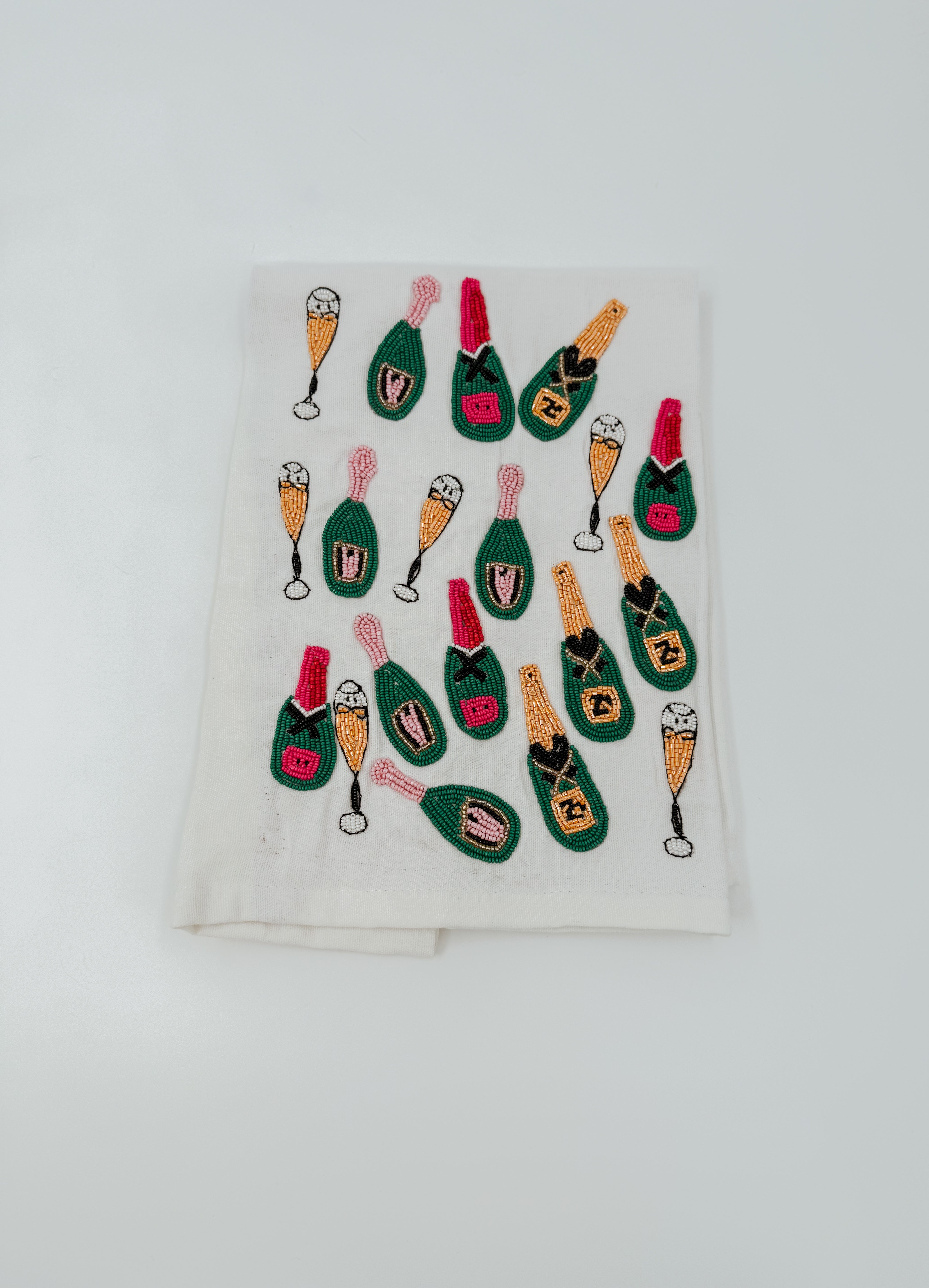 An off-white fabric napkin featuring a repeating pattern of embroidered champagne bottles and champagne flutes in green, pink, orange, and black.