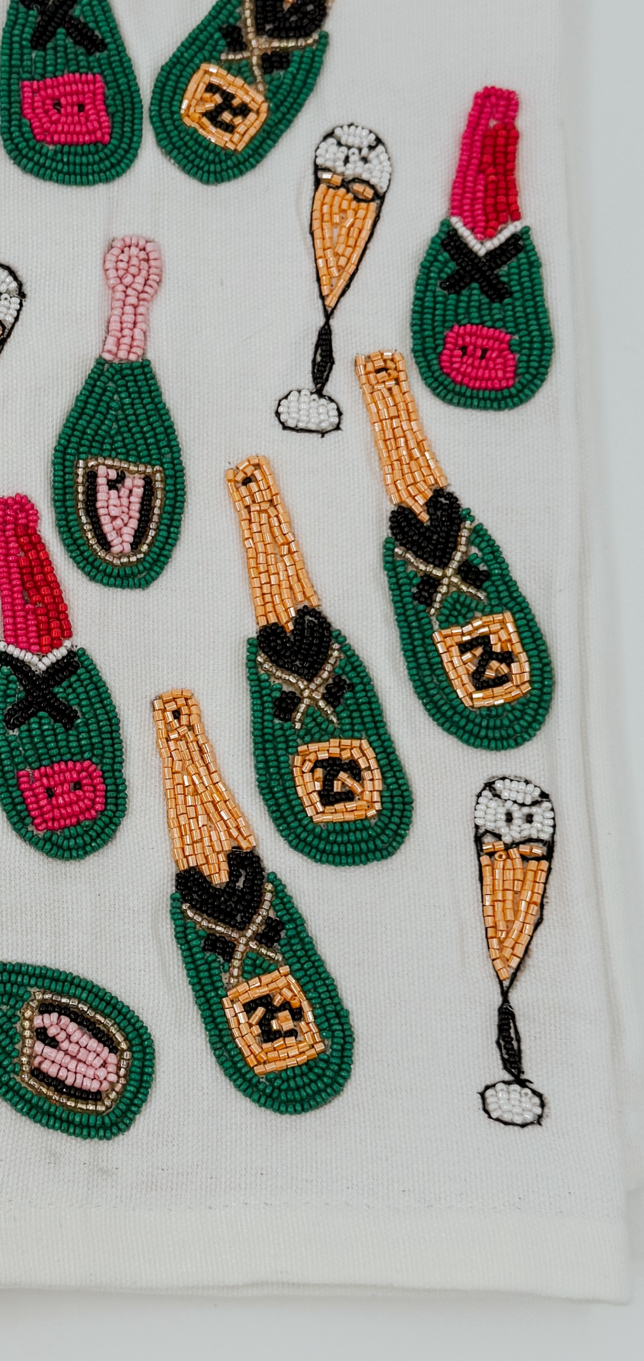 A close-up of a white textile featuring embroidered champagne bottle and champagne flute motifs in green, gold, black, pink, and white.